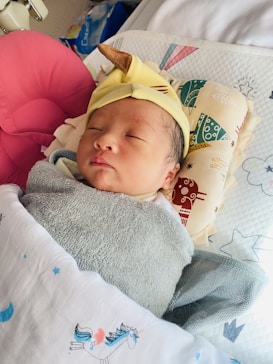 A newborn baby is peacefully sleeping, wrapped in a soft gray blanket and covered with a white blanket featuring unicorn and star prints. The baby is wearing a yellow hat with a cute brown horn, and rests on a patterned pillow.