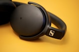 A pair of over-ear headphones with a sleek black design rests on a smooth surface. The ear cups are large and round, connecting to a comfortable headband. The headphones feature a prominent logo on the side, displaying a sense of premium quality and modern design.