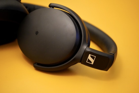 A pair of over-ear headphones with a sleek black design rests on a smooth surface. The ear cups are large and round, connecting to a comfortable headband. The headphones feature a prominent logo on the side, displaying a sense of premium quality and modern design.