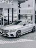 A sleek silver Mercedes-Benz car is parked on a cobbled driveway in front of a glass-front dealership. The reflection in the windows shows nearby buildings and part of the street. A woman stands near the entrance looking at her phone.