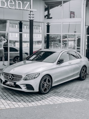 A sleek silver Mercedes-Benz car is parked on a cobbled driveway in front of a glass-front dealership. The reflection in the windows shows nearby buildings and part of the street. A woman stands near the entrance looking at her phone.