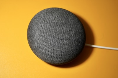 A small, round, fabric-covered smart speaker is placed on a bright yellow surface, with a white cable attached.