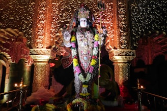 A detailed statue adorned with vibrant garlands stands in an ornately decorated interior with elaborate carvings and golden tones. The statue is centered and appears to depict a deity holding a trident. Two small lamps with flames are placed symmetrically in the foreground.