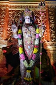 An intricately decorated statue of a deity adorned with vibrant floral garlands and elaborate jewelry. The sculpture is set against a richly carved golden background, holding a trident in one hand while the other hand is raised in a gesture of blessing.