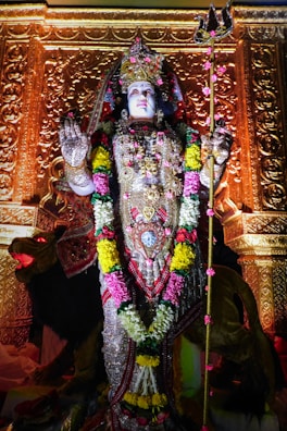 An intricately decorated statue of a deity adorned with vibrant floral garlands and elaborate jewelry. The sculpture is set against a richly carved golden background, holding a trident in one hand while the other hand is raised in a gesture of blessing.