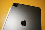 A sleek, metallic tablet with an Apple logo on the back sits on a yellow surface. Next to it, a white stylus is aligned parallel to the device.
