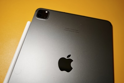A sleek, metallic tablet with an Apple logo on the back sits on a yellow surface. Next to it, a white stylus is aligned parallel to the device.