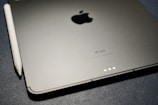 A close-up view of an Apple iPad and an Apple Pencil on a textured dark surface. The iPad is positioned with the Apple logo and the word 'iPad' visible. The Apple Pencil is placed alongside the iPad.