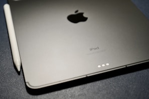 A close-up view of an Apple iPad and an Apple Pencil on a textured dark surface. The iPad is positioned with the Apple logo and the word 'iPad' visible. The Apple Pencil is placed alongside the iPad.