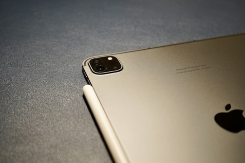 A close-up view of an Apple device, likely an iPad, paired with a stylus resting on its surface. The rear camera is visible, along with the Apple logo. The background is a textured gray surface.