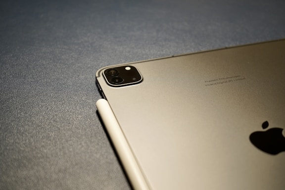 A close-up view of an Apple device, likely an iPad, paired with a stylus resting on its surface. The rear camera is visible, along with the Apple logo. The background is a textured gray surface.