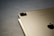 A close-up view of an Apple device, likely an iPad, paired with a stylus resting on its surface. The rear camera is visible, along with the Apple logo. The background is a textured gray surface.