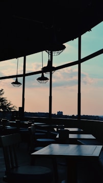 A dimly lit cafe or restaurant interior with empty tables and chairs is viewed against a backdrop of large windows, through which an evening sky with scattered clouds is visible. The silhouette of structural beams and ceiling lights is evident, adding a modern and slightly industrial feel to the setting.