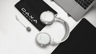 A pair of white headphones with a detachable microphone is placed on a white surface beside a black pouch labeled 'DAXA Series.' Nearby, a portion of a laptop is visible along with a black mouse pad.