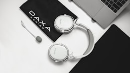 A pair of white headphones with a detachable microphone is placed on a white surface beside a black pouch labeled 'DAXA Series.' Nearby, a portion of a laptop is visible along with a black mouse pad.