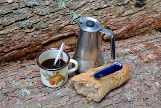 A stovetop coffee maker, a ceramic mug with a floral design containing coffee and a spoon, and a loaf of rustic bread with a pocket knife on top are placed on a textured wooden log surface.