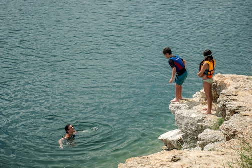 Three individuals are engaged in a recreational activity by a body of water. One person is swimming and looking up from the water with a smile, while two others, one boy and one girl, both wearing life jackets, stand on a rocky ledge, possibly preparing to jump into the water.