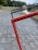 A close-up view of a red metal frame intersecting with a black one, positioned diagonally against a paved walkway. The background features blurred greenery and structures, suggesting an outdoor setting.