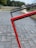 A close-up view of a red metal frame intersecting with a black one, positioned diagonally against a paved walkway. The background features blurred greenery and structures, suggesting an outdoor setting.