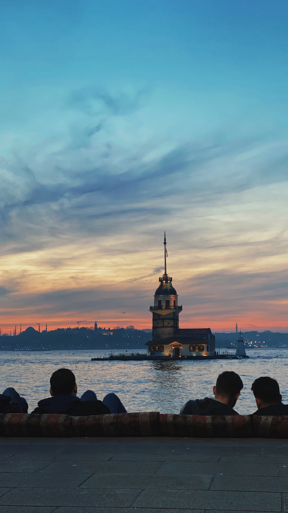 Group of people sitting on bench overlooking Bosphorus strait at sunset with bridge in background