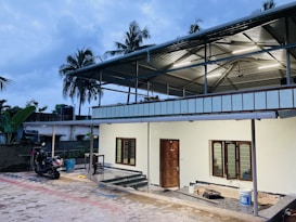 A modern building with a metal roof and a small porch area. There are two windows with grills beside a wooden door. A scooter is parked on a paved area in front of the building. The surrounding area includes palm trees and a slightly overcast sky.