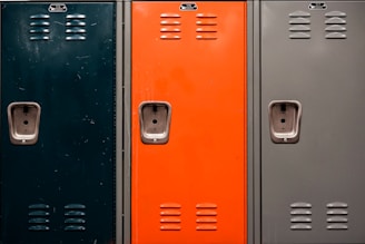 Three metal lockers side by side, each with a different color: dark green, orange, and grey. Each locker has a number plate, ventilation slots, and a recessed handle with a lock mechanism.