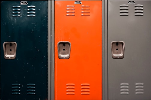 Three metal lockers side by side, each with a different color: dark green, orange, and grey. Each locker has a number plate, ventilation slots, and a recessed handle with a lock mechanism.