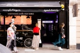 A group of people is seen walking past a storefront with signage promoting organic and healthy food options. Some individuals are wearing masks, indicating a concern for health or a response to current public health guidelines. A bicycle is parked in front of the store, and the overall ambiance is casual and modern.