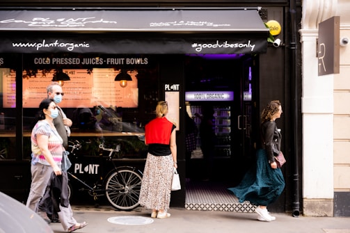 A group of people is seen walking past a storefront with signage promoting organic and healthy food options. Some individuals are wearing masks, indicating a concern for health or a response to current public health guidelines. A bicycle is parked in front of the store, and the overall ambiance is casual and modern.