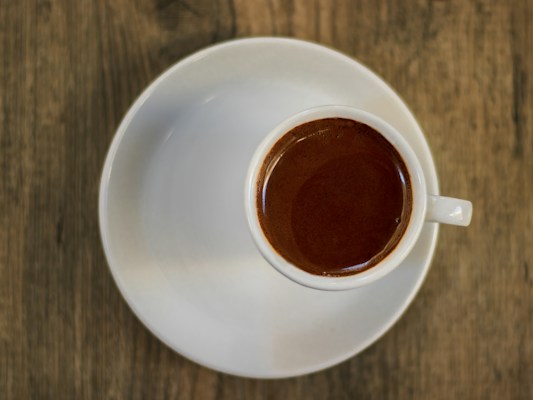A small white ceramic cup filled with a dark, rich espresso sits atop a matching white saucer on a wooden table. The warm, earthy tones of the wood contrast with the bright white of the cup, highlighting the deep brown color of the coffee.