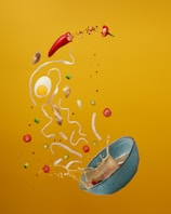 A dynamic composition of various food ingredients against a vibrant yellow background. The scene includes a cracked egg, slices of red chili peppers, pieces of ginger, noodles, and green onions, all seemingly suspended mid-air. A blue bowl appears to be tipping over, spilling its contents and creating an impression of motion.