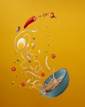 A dynamic composition of various food ingredients against a vibrant yellow background. The scene includes a cracked egg, slices of red chili peppers, pieces of ginger, noodles, and green onions, all seemingly suspended mid-air. A blue bowl appears to be tipping over, spilling its contents and creating an impression of motion.