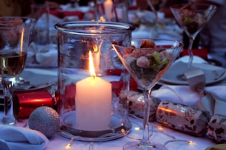 A dining table set for a festive or formal occasion features a large candle in a glass holder as the centerpiece, surrounded by glasses of wine and martini glasses filled with shrimp cocktail. Decorative elements include Christmas crackers and a shiny silver ornament. Soft lighting enhances the ambiance.