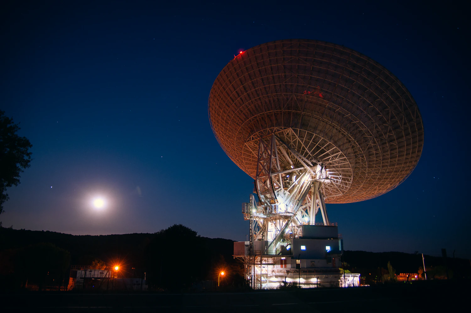 A giant radio dish lit orange at night with the moon rising behind it
