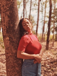 woman in red shirt and blue denim shorts leaning on brown tree