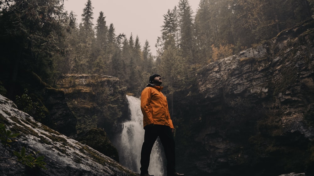 man in brown jacket standing on rock near waterfalls during daytime