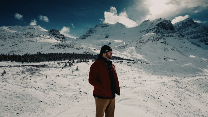 Bearded man in a red quilted insulated jacket and dark beanie standing on a snowfield below rugged snow-covered mountains