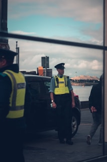 A police officer wearing a high-visibility vest stands near a parked car by the waterfront, with a cloudy sky and a view of the sea in the background. Part of another officer can be seen on the left, and a person walking away on the right. The scene appears to be captured through a reflective surface.