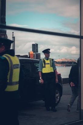 A police officer wearing a high-visibility vest stands near a parked car by the waterfront, with a cloudy sky and a view of the sea in the background. Part of another officer can be seen on the left, and a person walking away on the right. The scene appears to be captured through a reflective surface.