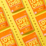 Several vibrant orange snack packages with the label 'LOVE CORN' are arranged diagonally on a bright yellow background. Each package is labeled as 'vegan cheesy flavored' and features corn kernels scattered around them.