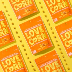 Several vibrant orange snack packages with the label 'LOVE CORN' are arranged diagonally on a bright yellow background. Each package is labeled as 'vegan cheesy flavored' and features corn kernels scattered around them.