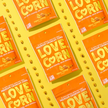 Several vibrant orange snack packages with the label 'LOVE CORN' are arranged diagonally on a bright yellow background. Each package is labeled as 'vegan cheesy flavored' and features corn kernels scattered around them.
