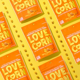 Several vibrant orange snack packages with the label 'LOVE CORN' are arranged diagonally on a bright yellow background. Each package is labeled as 'vegan cheesy flavored' and features corn kernels scattered around them.