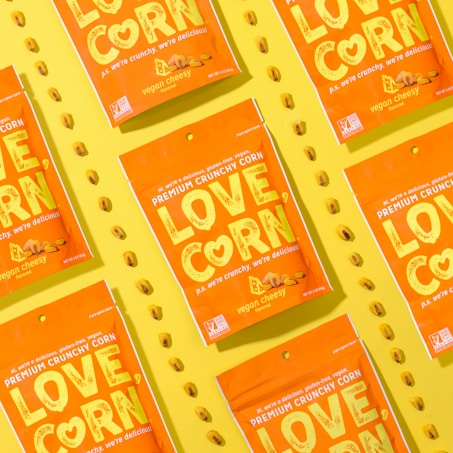 Several vibrant orange snack packages with the label 'LOVE CORN' are arranged diagonally on a bright yellow background. Each package is labeled as 'vegan cheesy flavored' and features corn kernels scattered around them.
