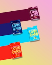 Four colorful packages of LOVE CORN, a premium crunchy corn snack, are arranged diagonally over a multicolored striped background. Each package represents a different flavor, distinguishable by their unique colors: purple, blue, teal, and red.