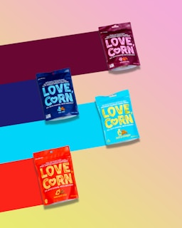 Four colorful packages of LOVE CORN, a premium crunchy corn snack, are arranged diagonally over a multicolored striped background. Each package represents a different flavor, distinguishable by their unique colors: purple, blue, teal, and red.