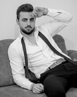 A man with a beard and styled hair is seated on a couch. He wears a white shirt, partially open with a tie hanging loosely around his neck, and black pants. His pose is relaxed with one arm resting on his forehead and the other by his side.