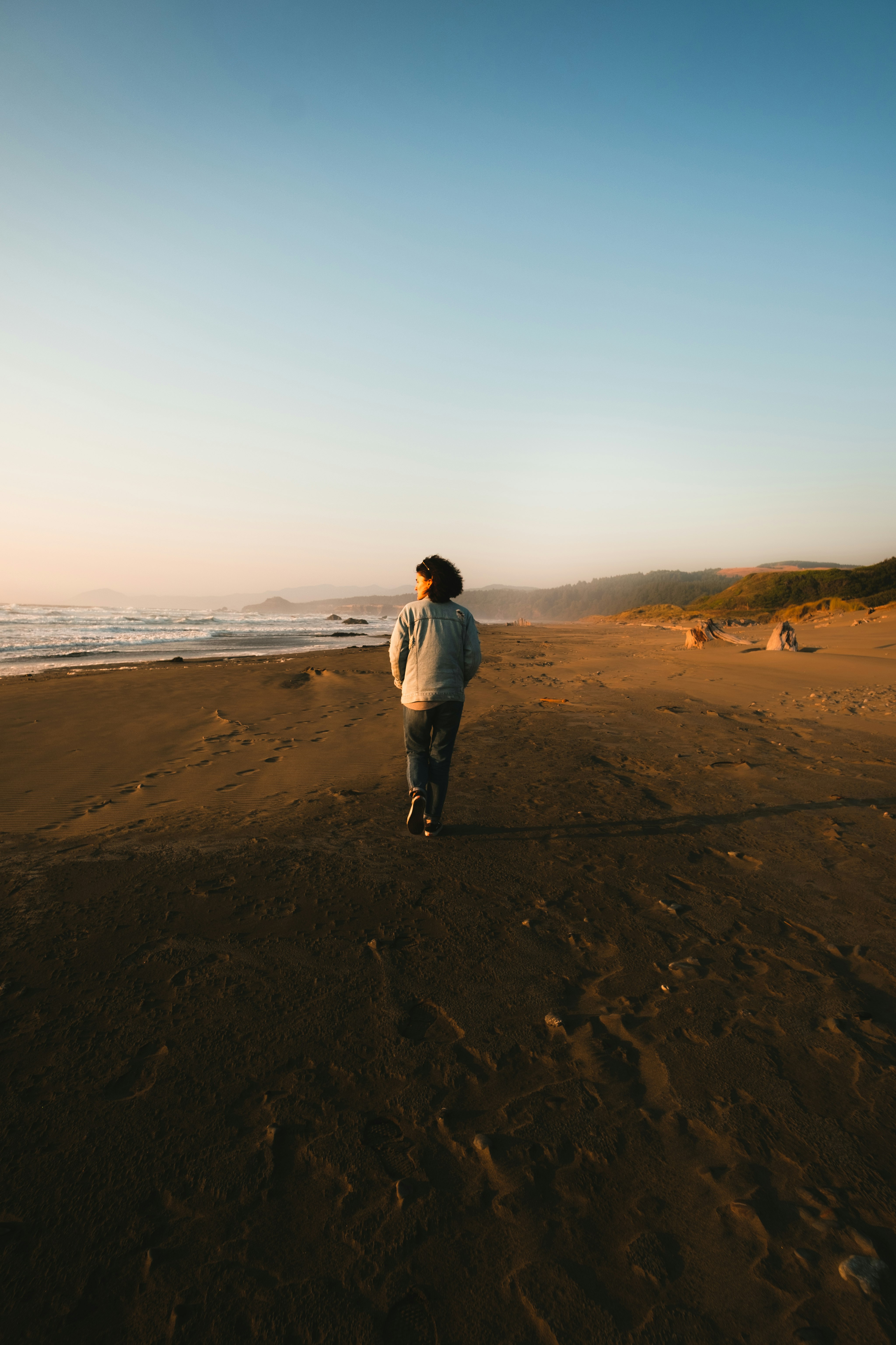 man in white jacket and blue denim jeans standing on brown sand near body of water