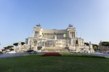 A grand neoclassical building with intricate carvings and statues, prominently featuring the Italian national flags. This impressive structure has large columns and is set in an urban area with trees and parked cars nearby. Visitors are seen at the base, giving a sense of its monumental scale.