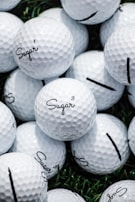 A collection of white golf balls with the word 'Sugar' printed on them in black script. The golf balls are stacked closely together on a surface with patches of green grass visible in the background.